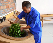 A man in matching blue smock and pants leaning over a slightly angled wok to expertly fry fresh rock wulong tea leaves by hand.