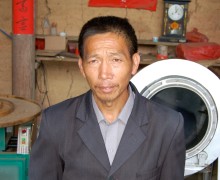 Tea Master Zhang Shuiquan standing in front of tea processing equipment, wearing a suit.