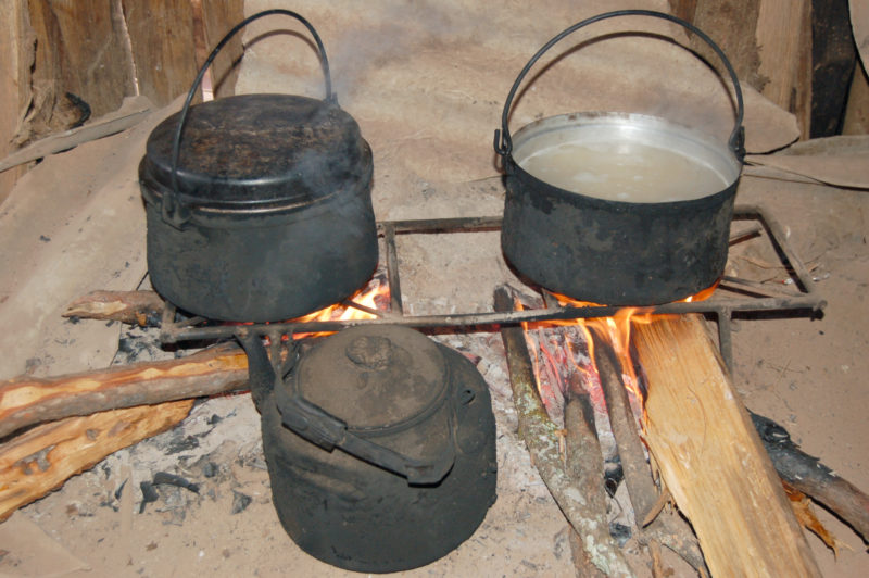 Two pots and a kettle boiling over a fire inside a village home.