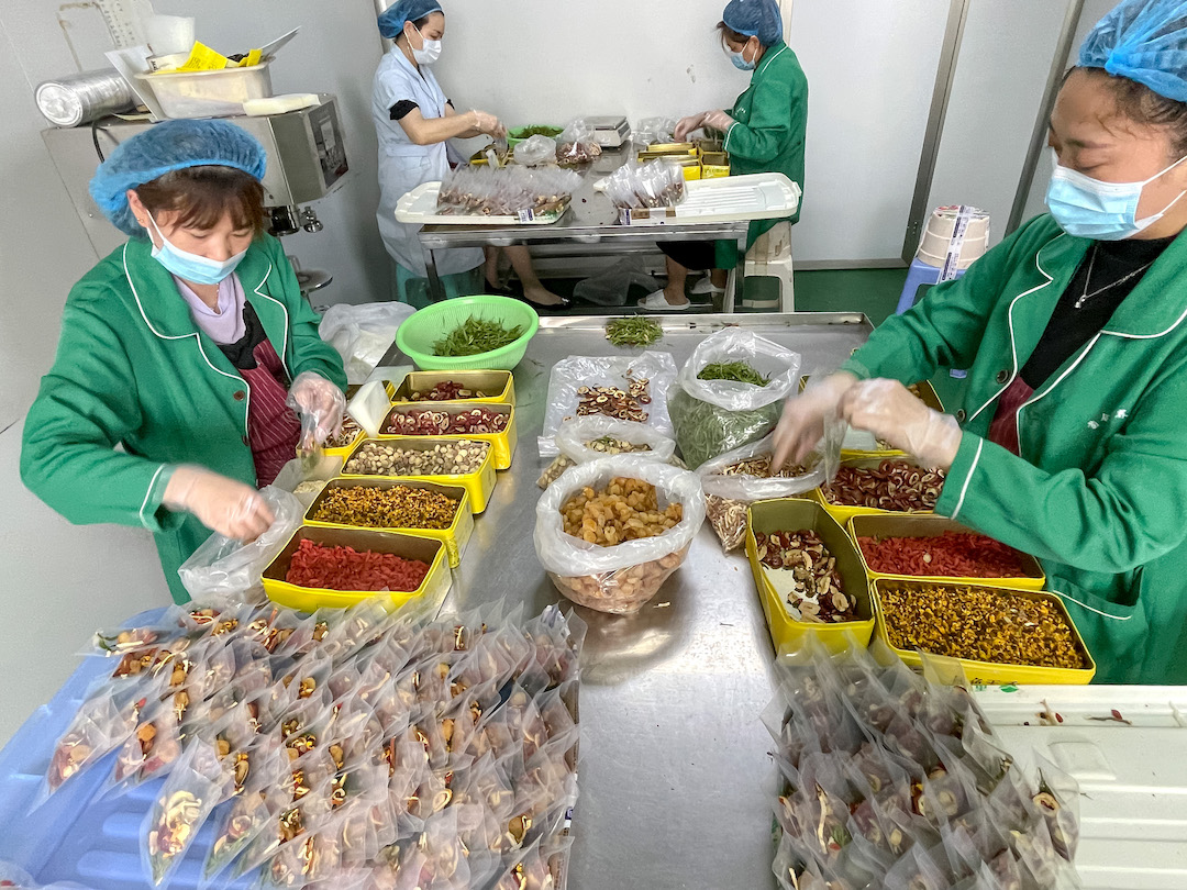 Four people wearing caps, gloves, and smocks set up at steel tables to assemble individual Eight Treasures tea packets from bags of several herbal ingredients.