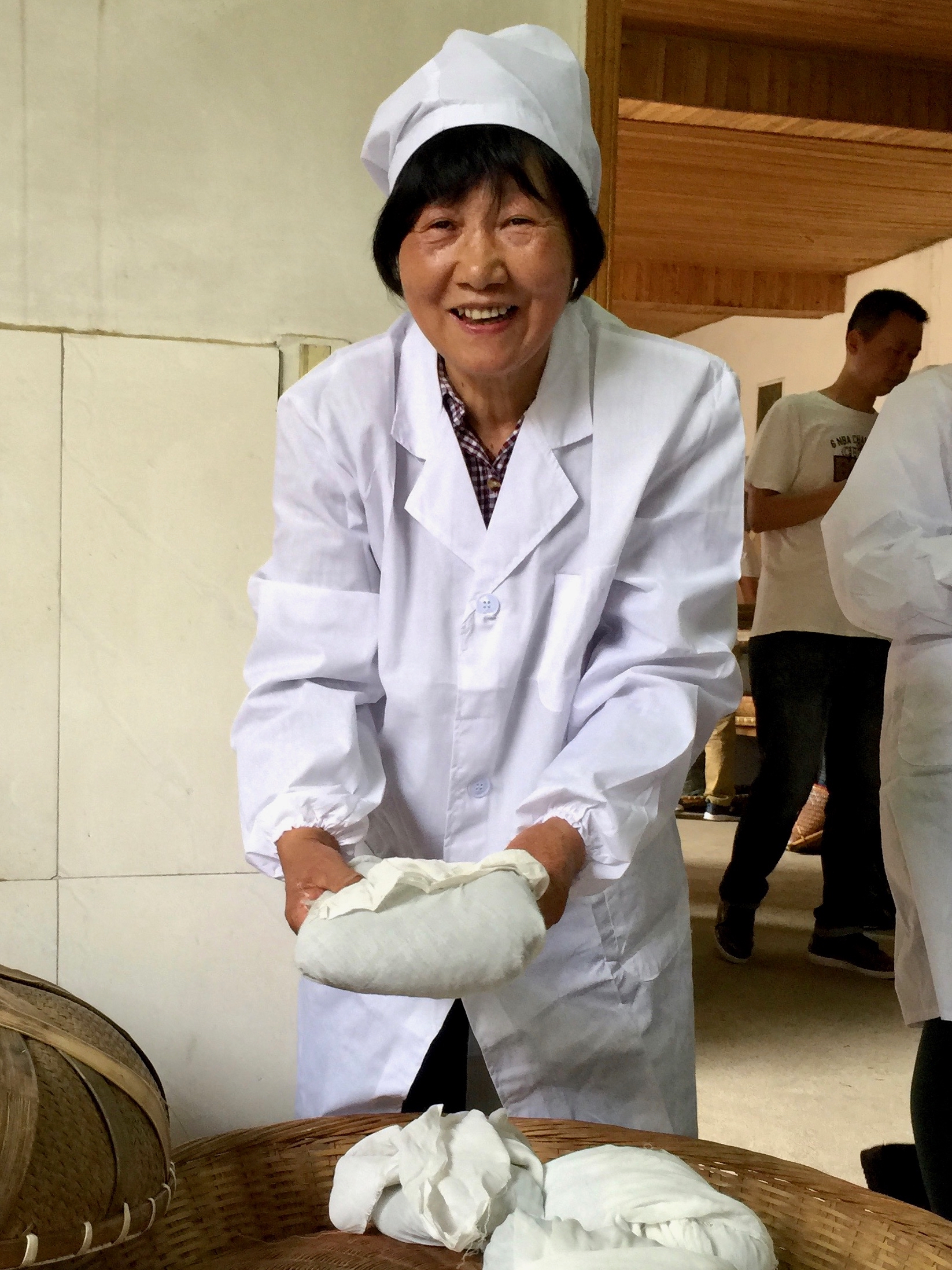 A smiling woman in a white coat and hat holding a small cloth-wrapped bundle in both hands.