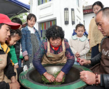 An older woman demonstrating frying Mogan tea by hand, surrounded by onlookers of all ages.
