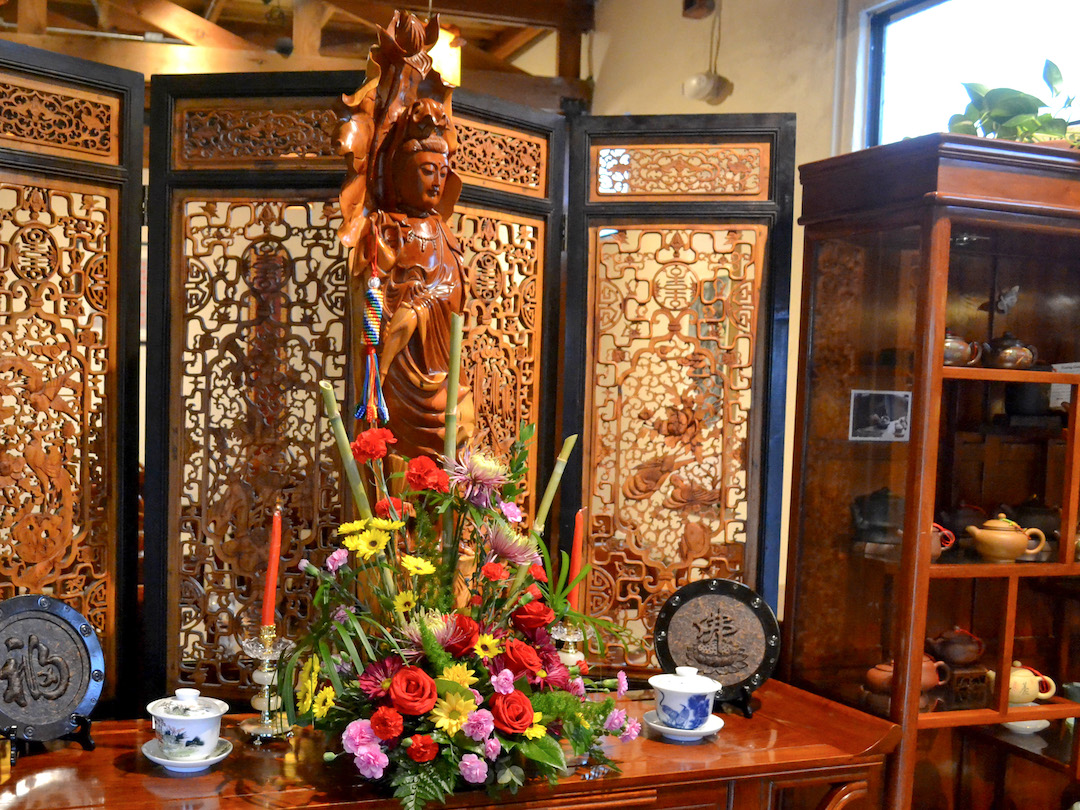 An altar indoors with a tall wooden statue of Guanyin surrounded by flowers, candles, and tea. Off to one side, a shelf of yixing clay teapots.