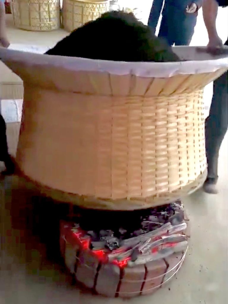 A large woven bamboo drum with Lu'an Gua Pian tea on top being lifted over an open charcoal brazier by two people.
