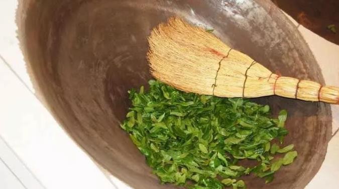 A 45° angled wok with a small batch of green tea leaves in it, being tossed and turned with a broom made of thin reeds.