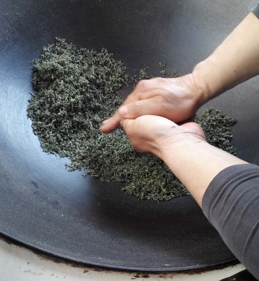 A pair of hand tightly pressing and rolling Bi Luo Chun tea leaves between them while frying in a large wok.