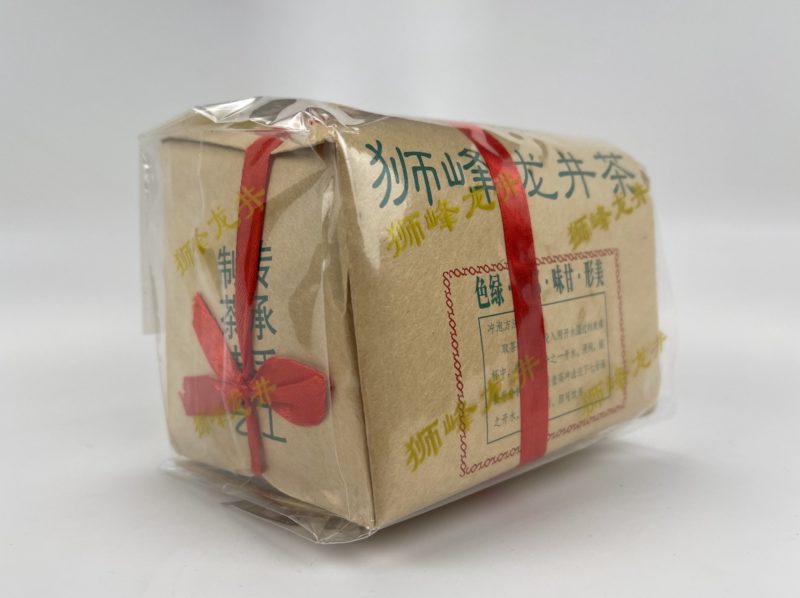 The traditional paper packaging of Shifeng Longjing printed with green Chinese characters showing the name of the tea. A red ribbon wraps the bundle.