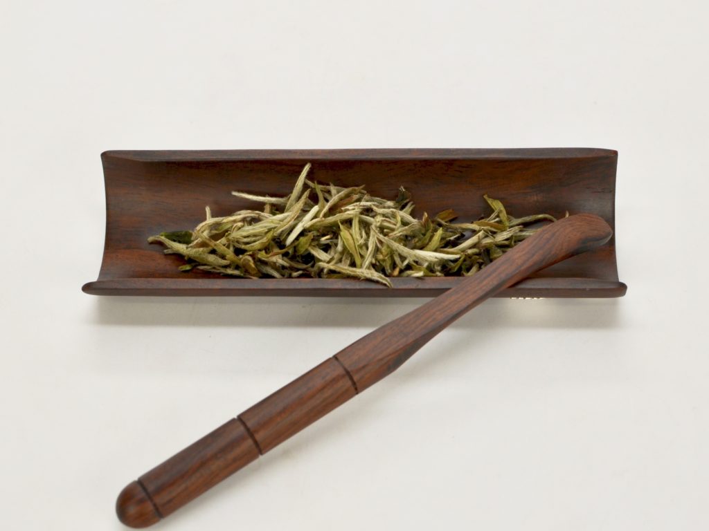 Dry Baihao Yinzhen (Silver Needle) white tea leaves on a wooden display plate.
