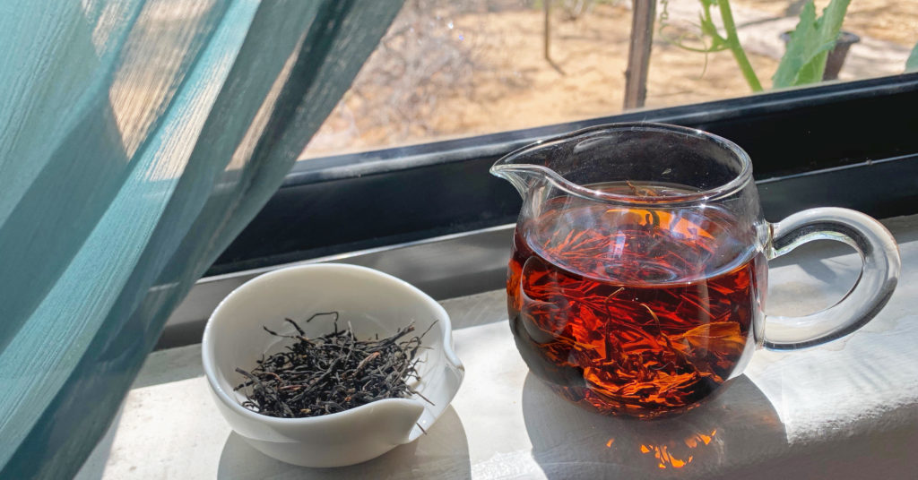 A pitcher of brewed black tea with a bright red infusion color, next to a dish filled with black dry tea leaves, perched in a windowsill with a blue curtain in the sun.