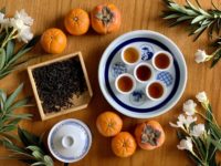 Five cups of amber rock wulong tea sit on a white and blue porcelain plate surrounded by dark tea leaves, orange fruits and stalks of white flowers.