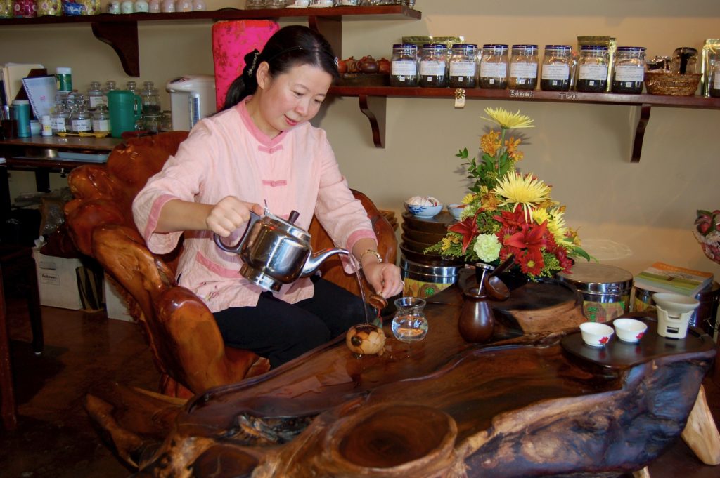 Zhuping pouring hot water from a kettle into a small yixing clay pot at a wooden tea table.