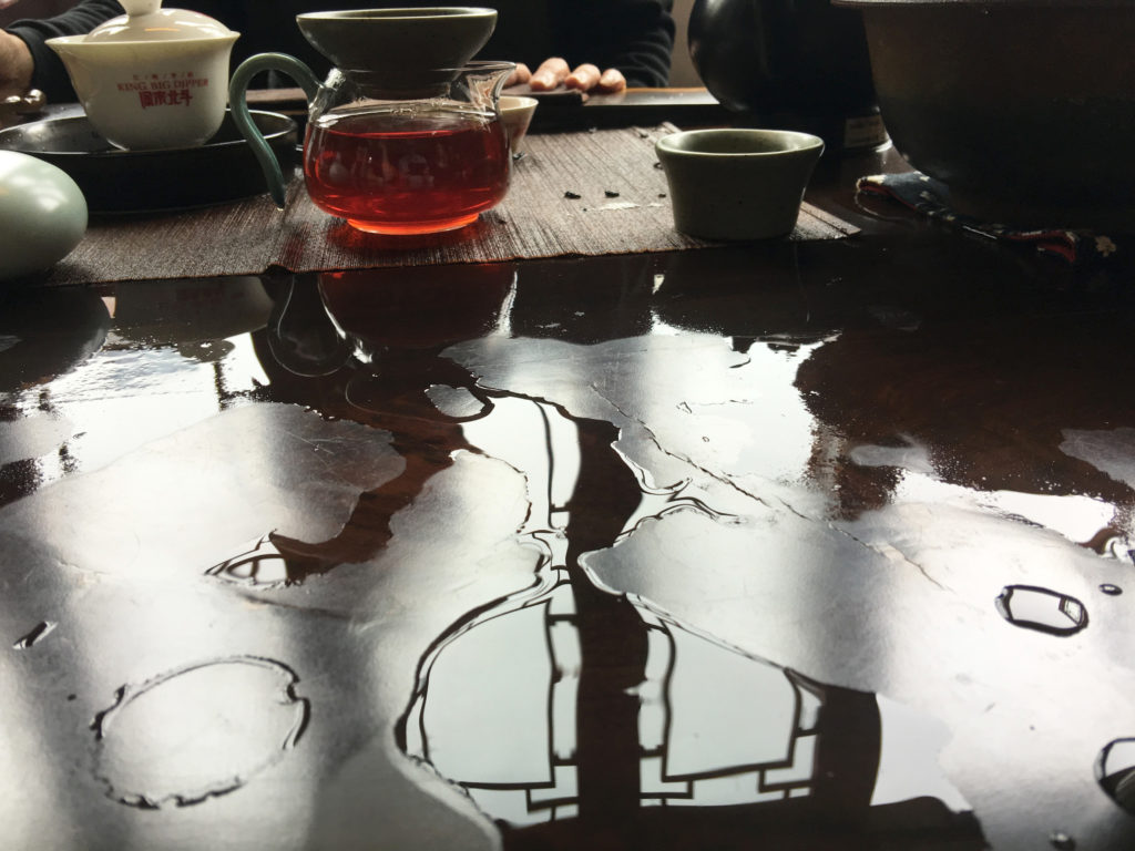 Spilled water reflecting light from the windows trailing across a tea table, with a full pitcher of deep red wulong tea and brewing accessories in the background.
