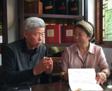 Chen Dehua and Zhuping Hodge happily conversing and gesturing, seated at a wooden table with teacups and notebooks on it, with shelves of ornate boxes of tea behind them.