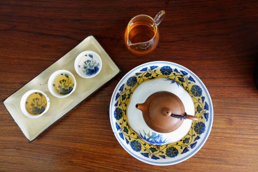 Above view of a small yixing clay teapot on a stand that holds it above spilled tea. Next to it is a pitcher of rock wulong tea and three small teacups on a long tray.