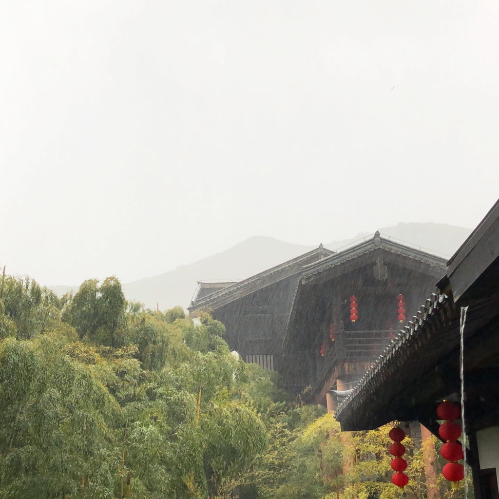 A hazy sky above bamboo forest and dark buildings with water running out the gutters, being drenched in pouring rain.
