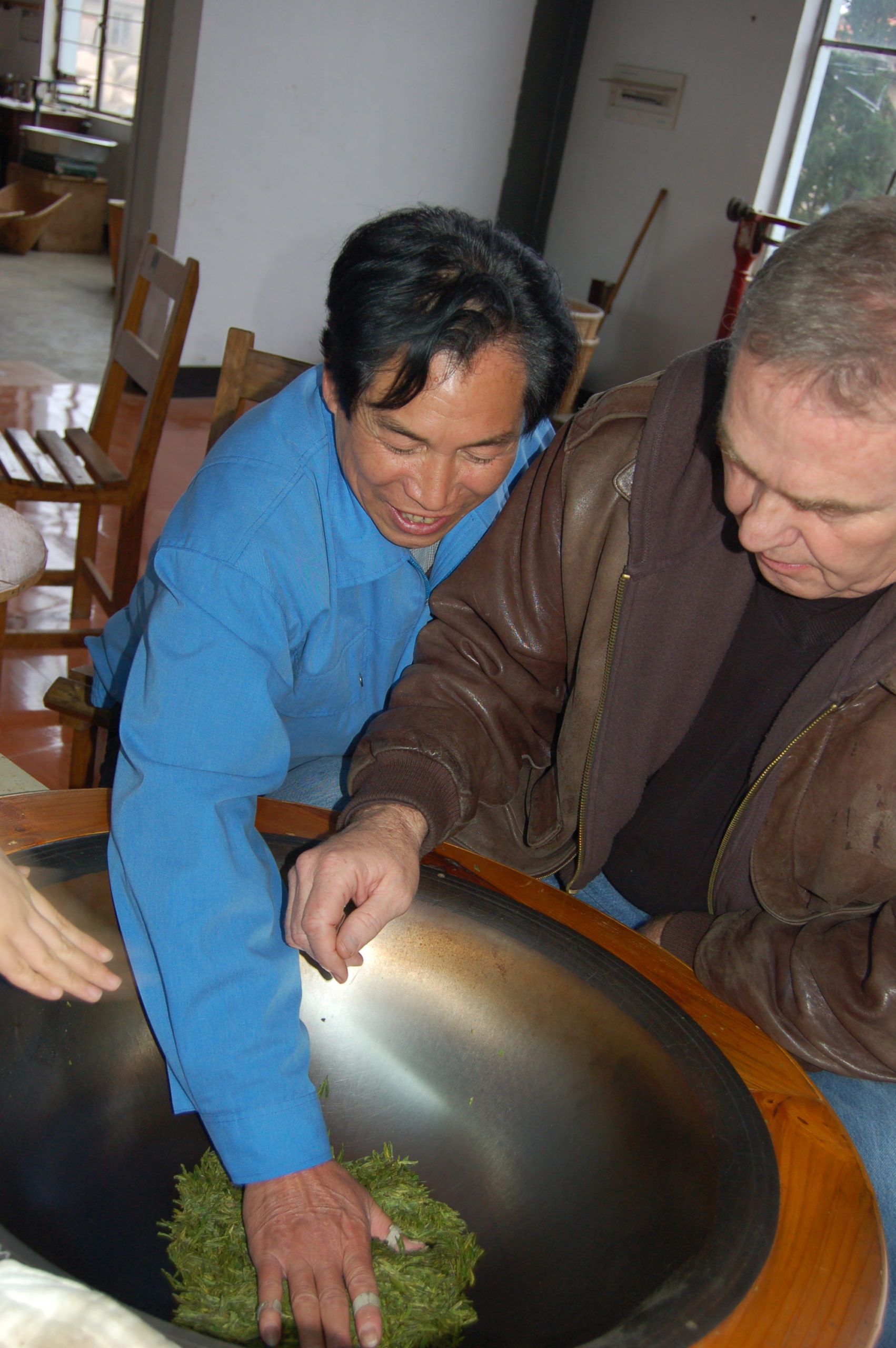 Two men standing over a wok with Longjing green tea leaves in it, one with a hand in the wok demonstrating and one learning.