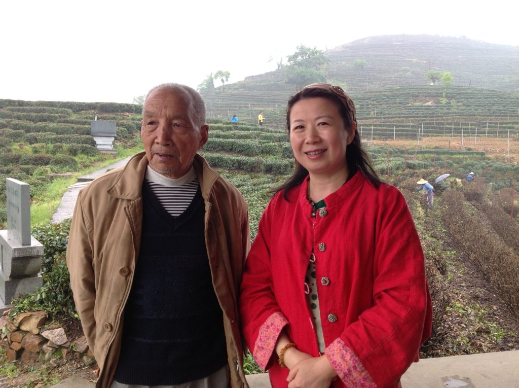 Mr. Weng and Zhuping standing side by side with low sloping misty hills covered in rows of tea bushes in the background. A few people among the rows are harvesting fresh leaves.