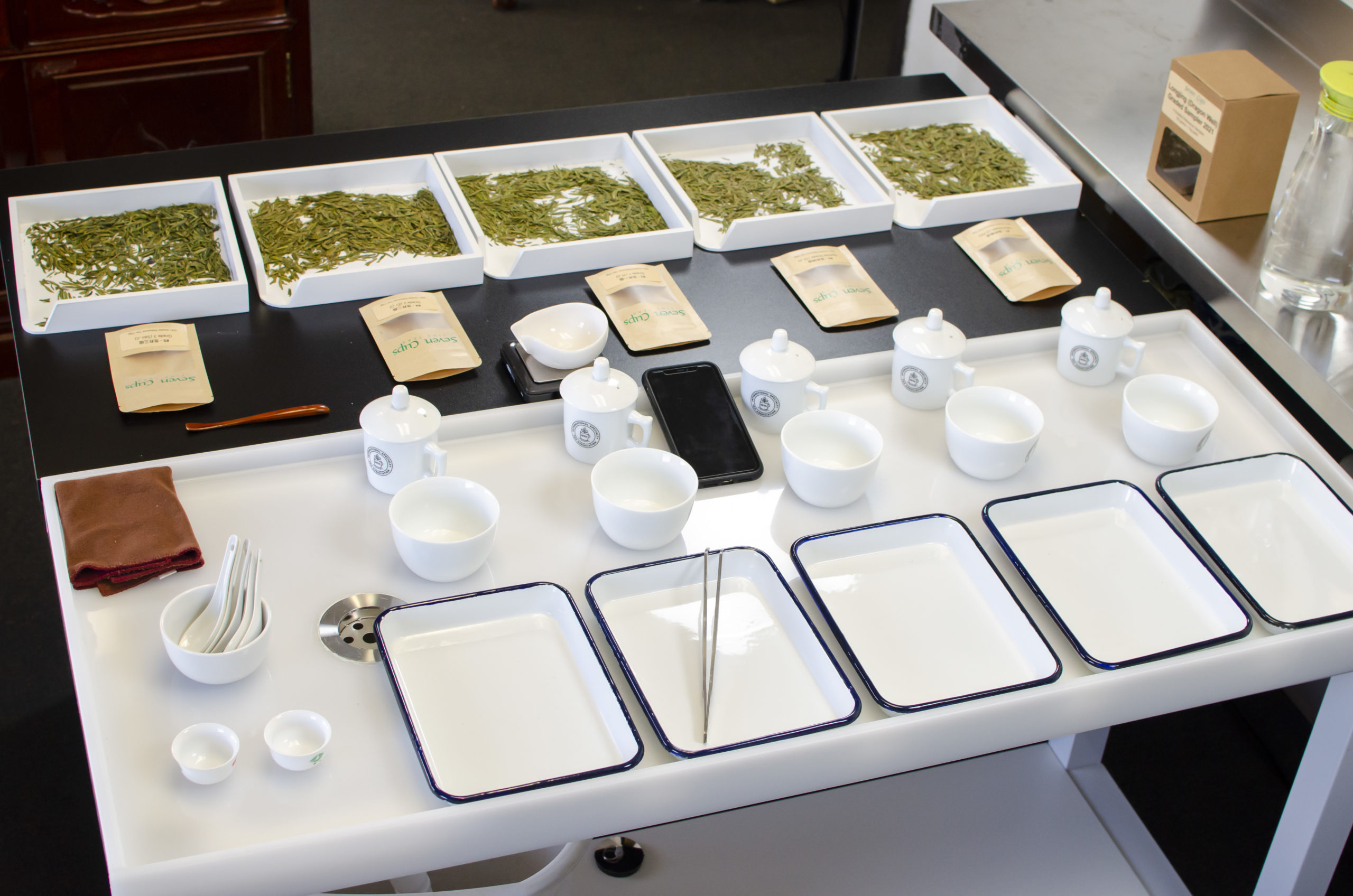 A tea judging setup spread out on a table, including an examination box for dry leaves, a brewing vessel, a bowl to pour tea into, and a tray for examining wet leaves for each of the five teas.
