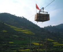 A cable line stretching over a mountainside covered in tea terraces in Huangshan, bearing a load of boxes marked with a red flag.