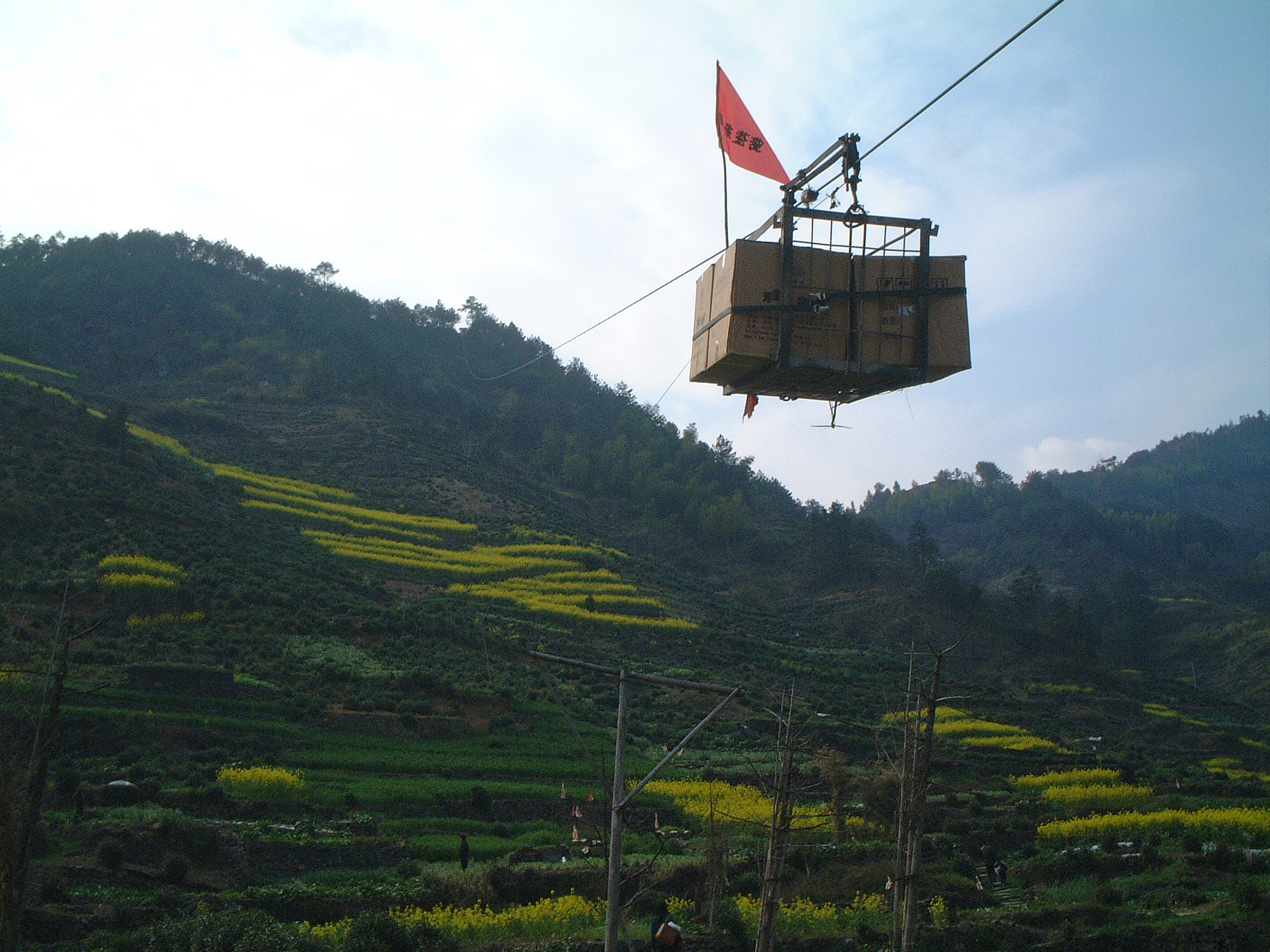 A cable line stretching over a mountainside covered in tea terraces in Huangshan, bearing a load of boxes marked with a red flag.