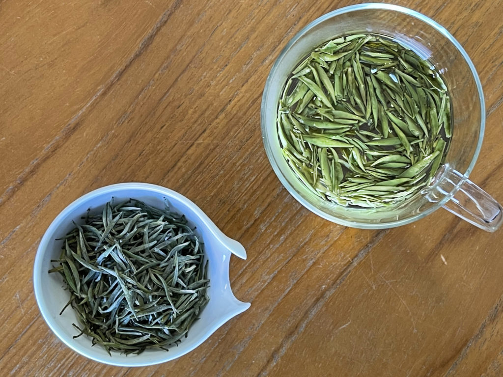 Looking down into a glass mug full of brewing needle-shaped tea leaves, next to a porcelain dish of the same dry leaves, both on a wooden surface.