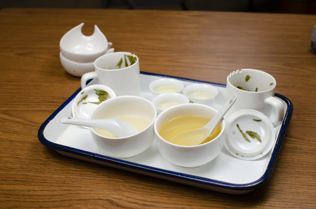 Two sets of white porcelain brewing equipment gathered on a tray on a wooden surface. Prominent in the front are two large bowls of infused golden-green tea.