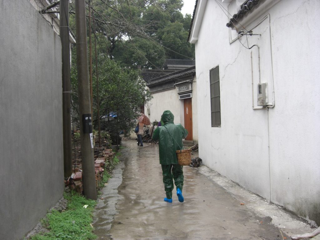 A tea plucker in a green rain coat walks down a narrow street between white buildings. The ground is wet with rain water. The figure carries a woven basket over one shoulder.