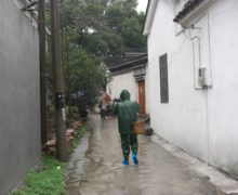 A tea plucker in a green rain coat walks down a narrow street between white buildings. The ground is wet with rain water. The figure carries a woven basket over one shoulder.