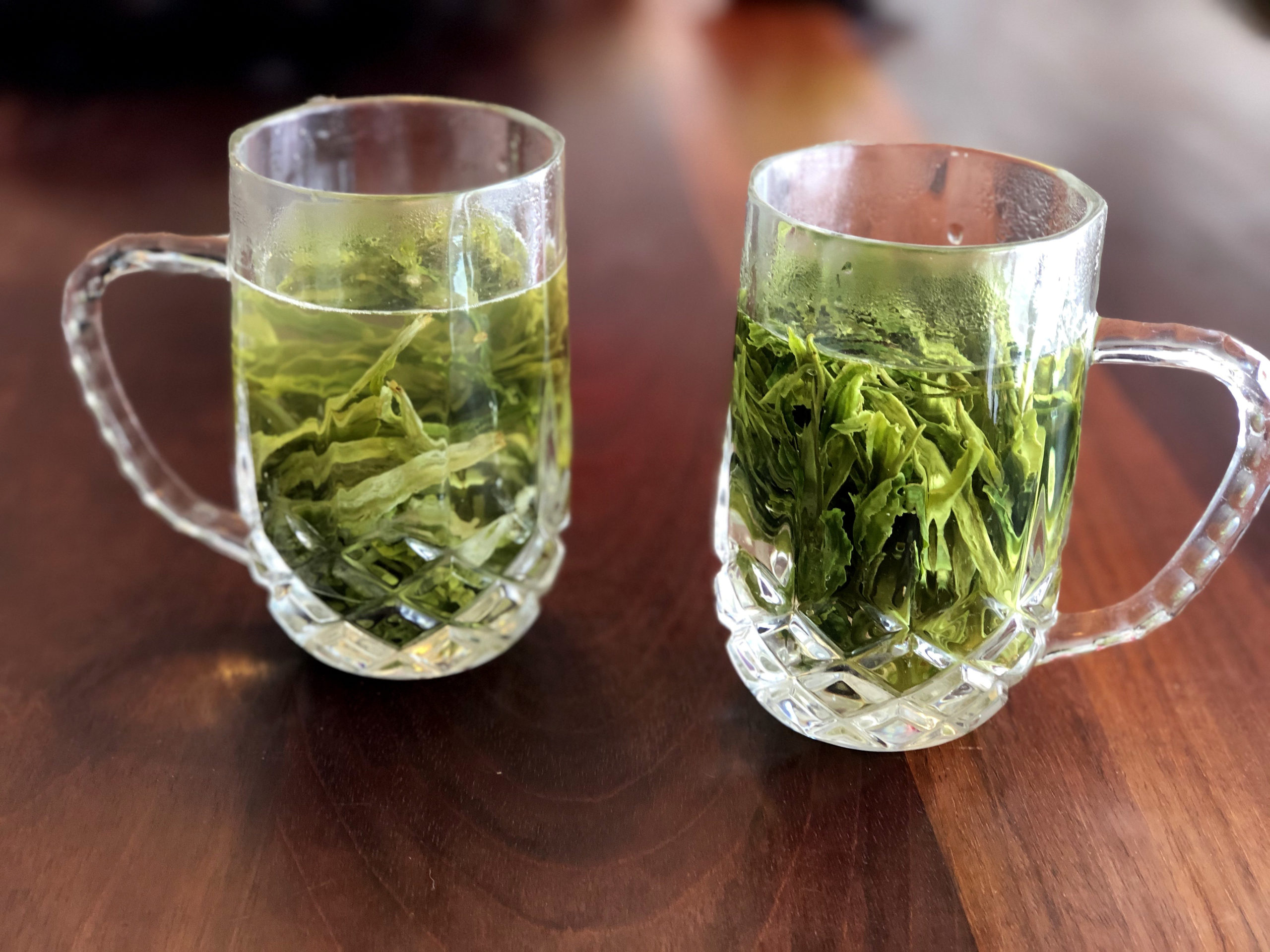 Two glass mugs sit on a wooden table. The mugs are filled with large green tea leaves steeping in hot water.