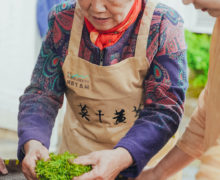 An older woman in an apron kneading tea leaves on a woven bamboo tray while a young woman holds the tray steady for her.
