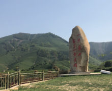 A tall monument of natural stone with red characters written on it. It stands on a small plinth near a fence, surrounded by the majestic low green mountains of Anji. A hand in the foreground points to the monument.