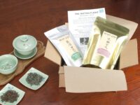 An open brown paper box with two bags of tea and an info sheet arranged inside for the Tasting Flight, accompanied by teaware set up for service on a tray.