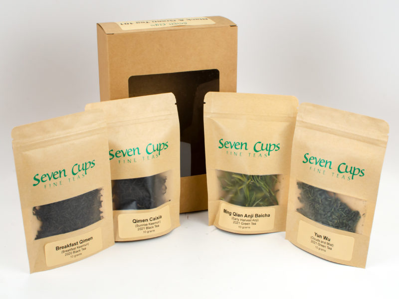 Small bags of the two green and two black teas included in the Black & Green Tea 101 sampler kit, arranged in front of its paper box.