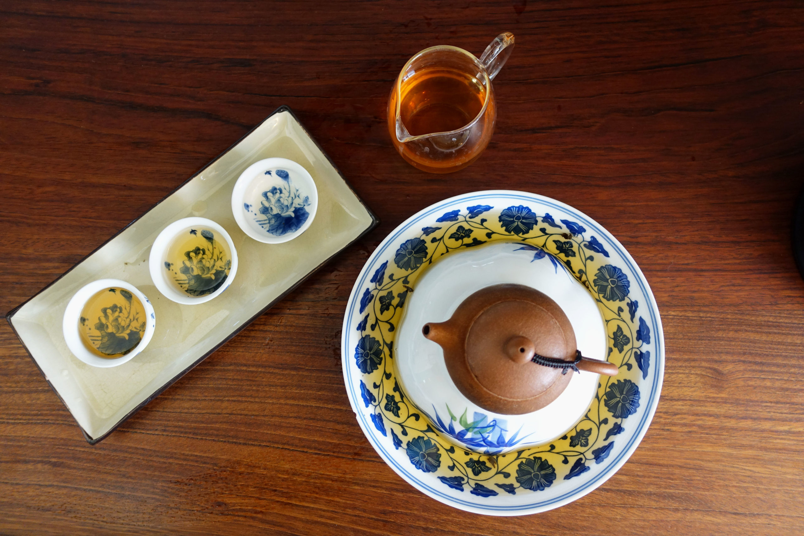 A speckled clay tea pot on a draining dish, with a pitcher full of tea and three tea cups next to it on a wooden table.