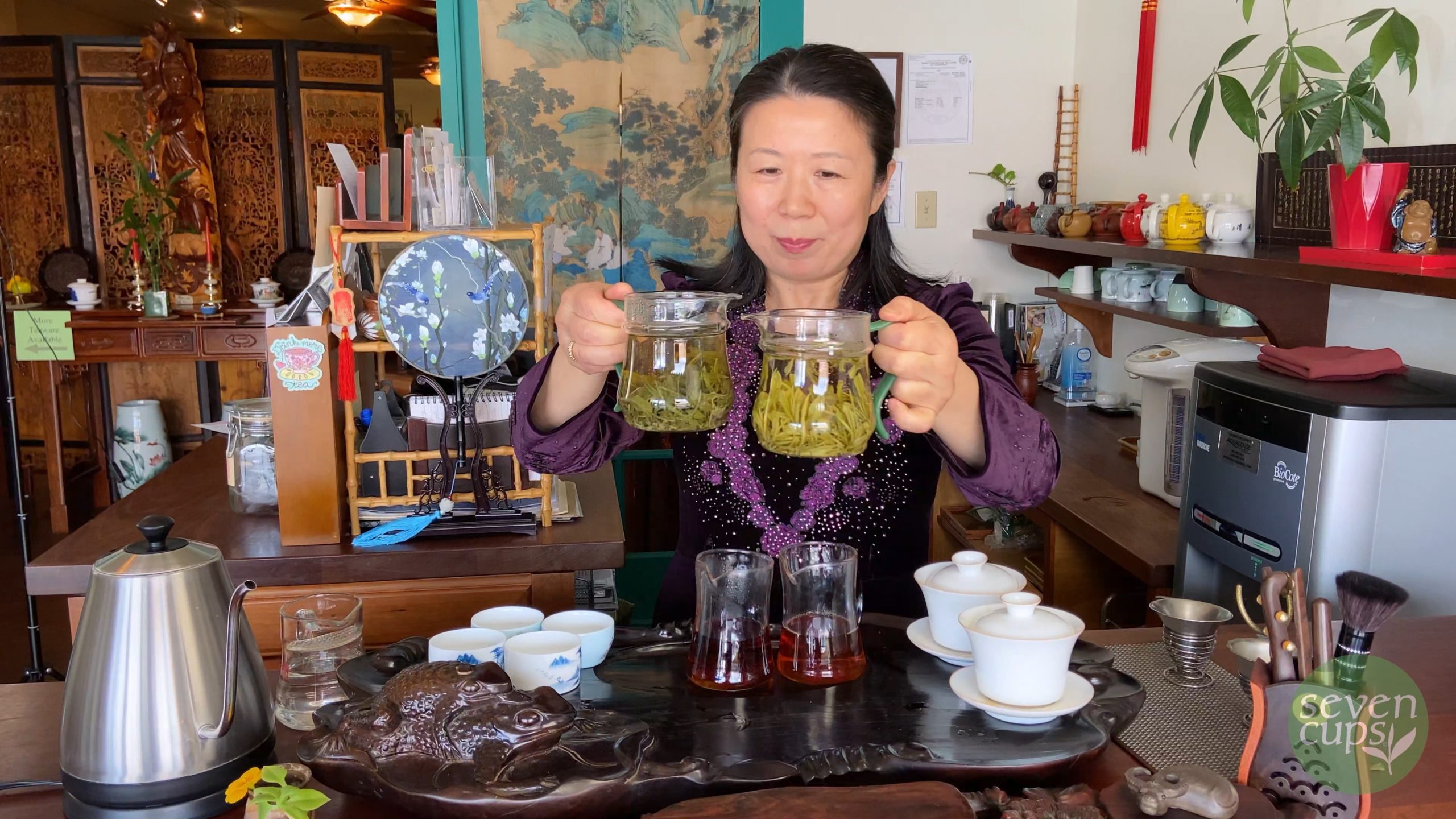 A woman standing in a traditional Chinese teahouse, smiling and holding up two glass pitchers filled with green tea side by side.