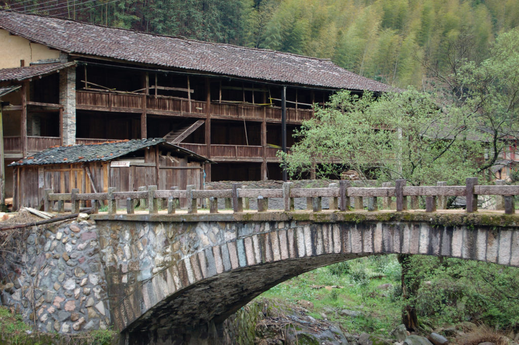 A stone bridge leading to an old building with three floors and open wooden balconies in a green forested valley.
