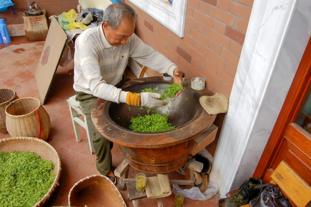 An elderly man with short grey hair expertly tossing tea leaves in a hot wok with both hands.