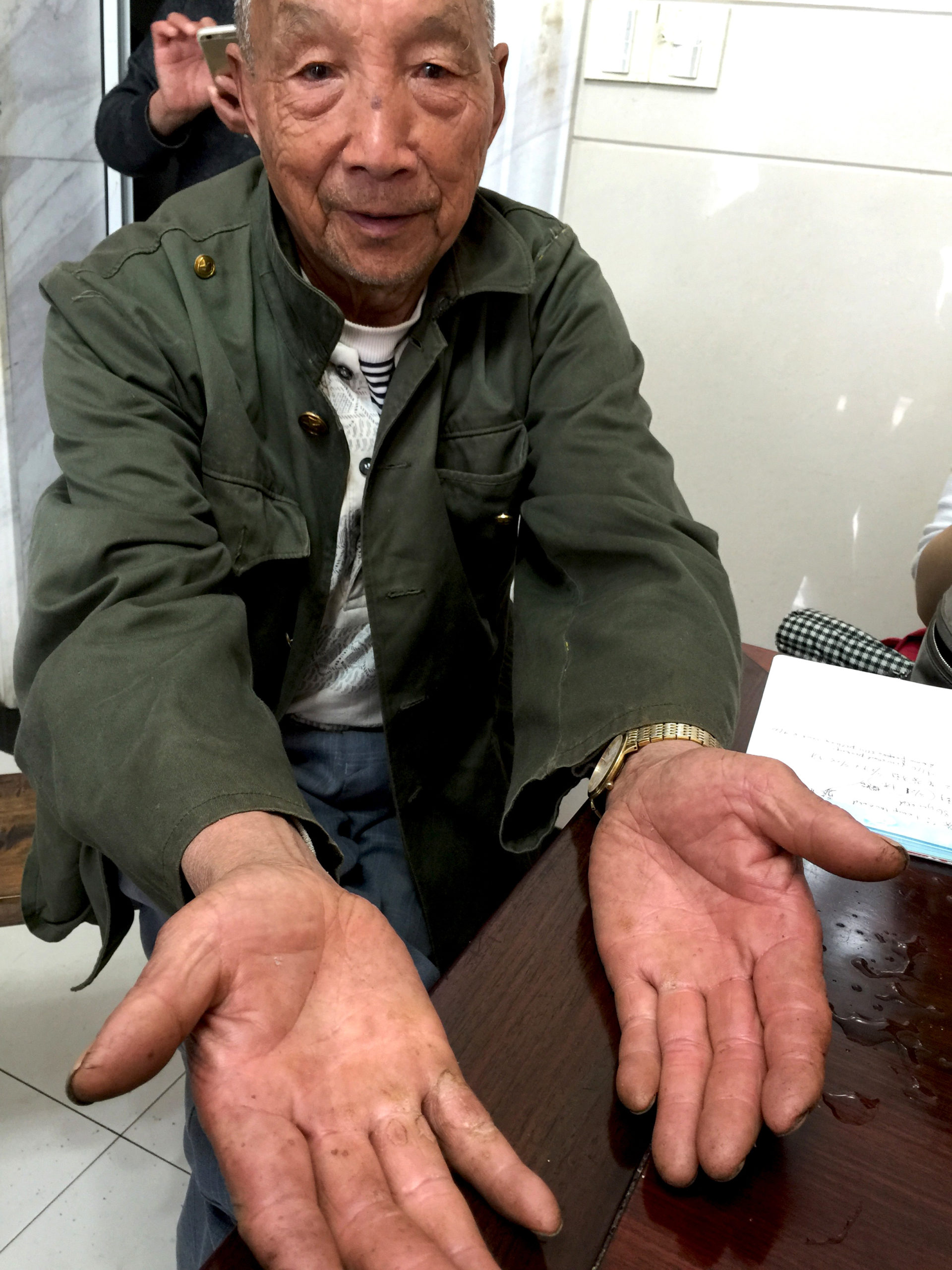 A faintly smiling elderly man extending his hands toward the viewer to show his upturned hands. The palms and fingers are thick with calluses, especially his right hand.