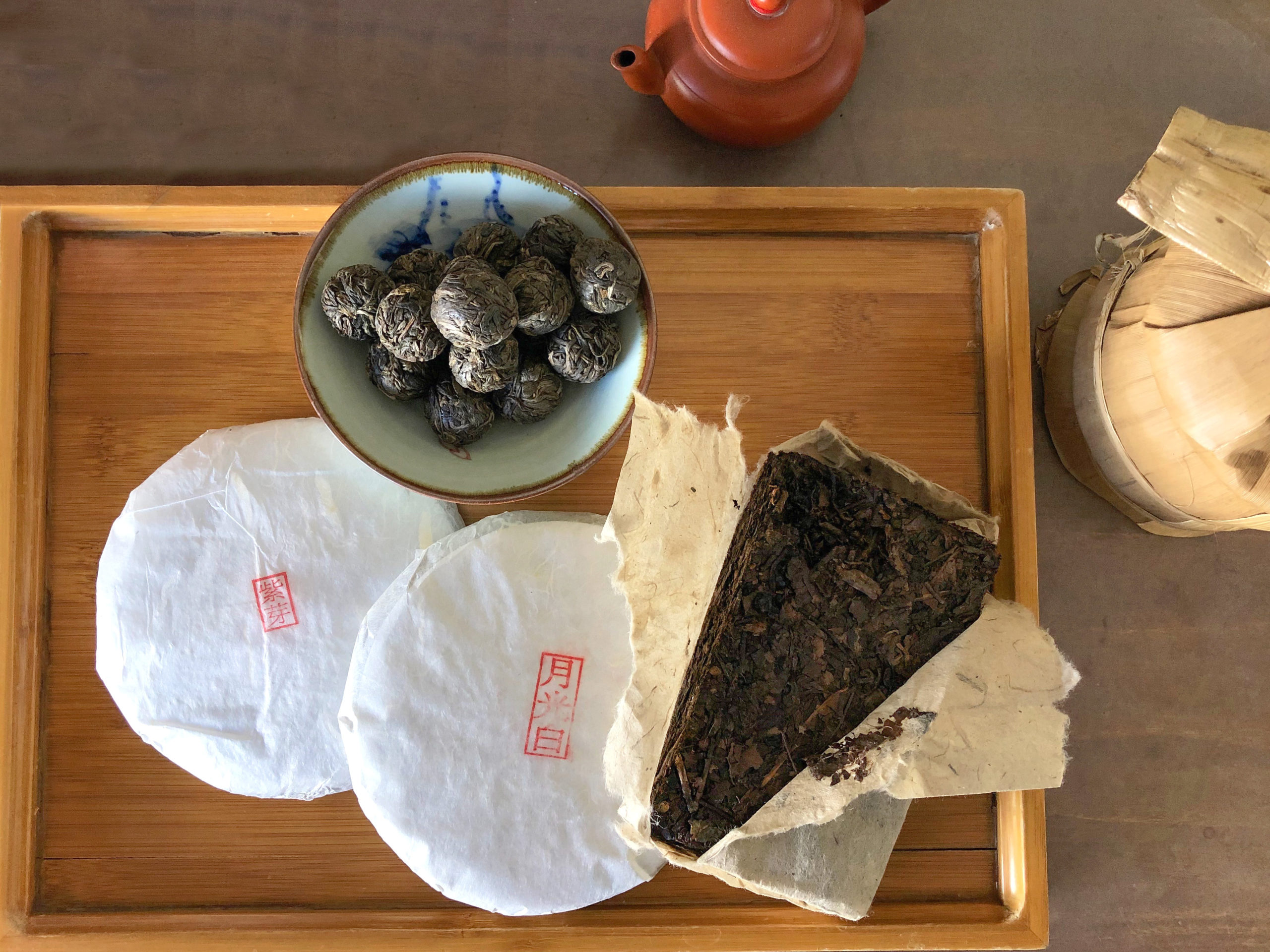 Several puer cakes on a bamboo tray, one with its paper wrapper opened to reveal the dark shu brick inside. A small clay tea pot and a bamboo-wrapped tong of more puer cakes sit on the table beside them.