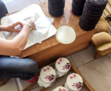 A person seated at a small workbench stacked high with finished small puer cakes, one by one wrapping them in paper and placing them on a wooden rack nearby.
