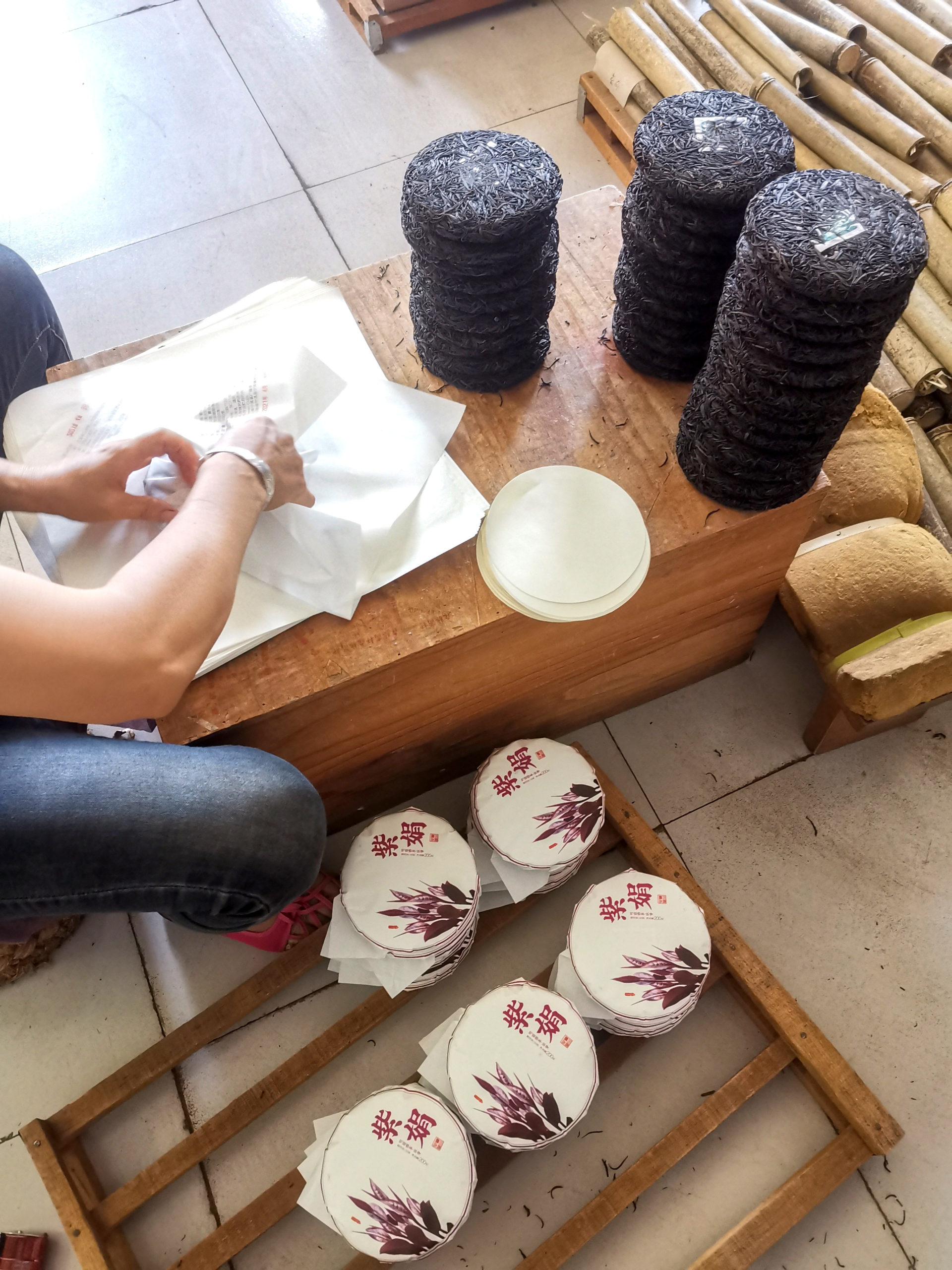 A person seated at a small workbench stacked high with finished small puer cakes, one by one wrapping them in paper and placing them on a wooden rack nearby.