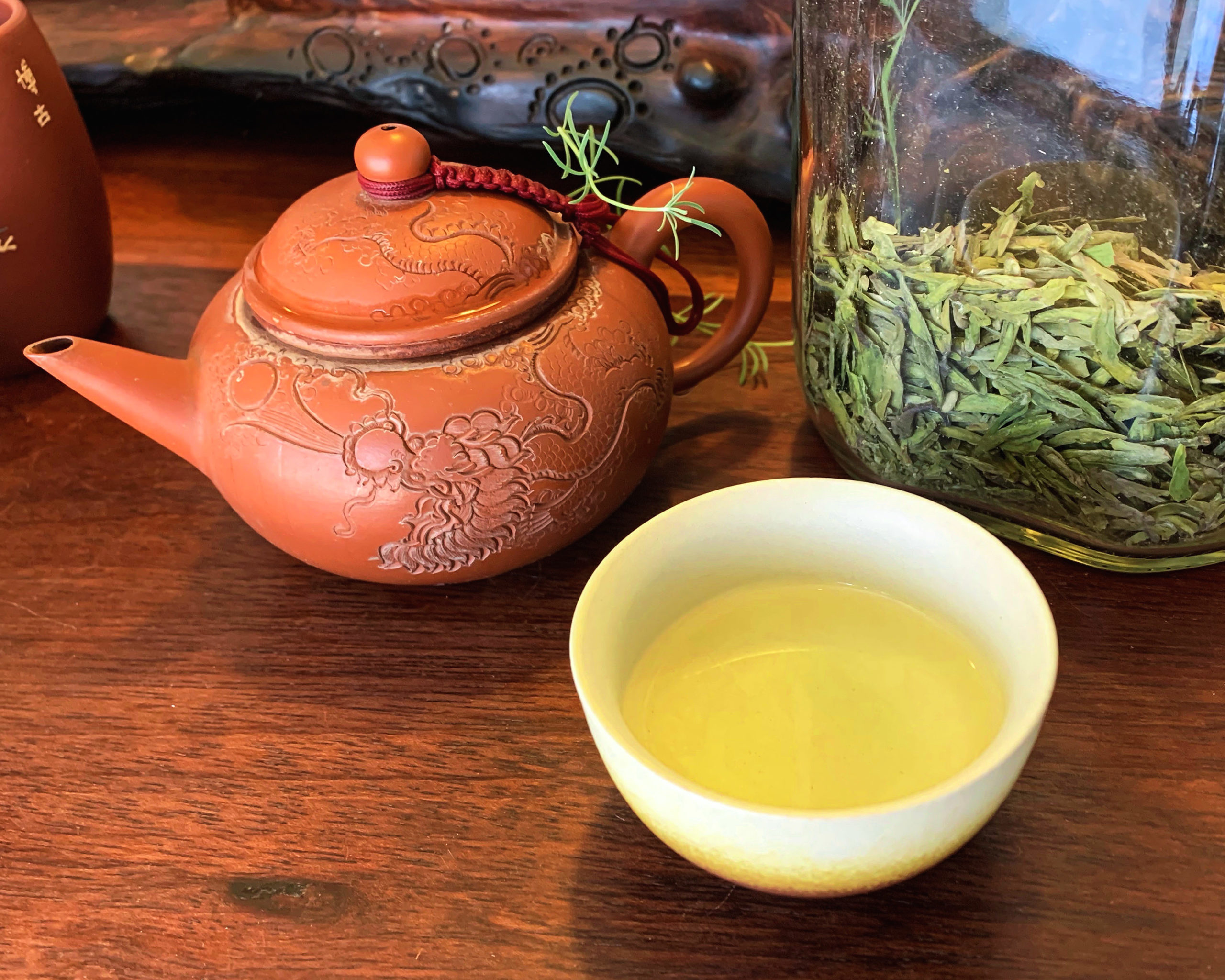 A red clay tea pot with a carved dragon pattern sitting on a wooden table next to a glass jar of dry Longjing tea and a small cup of brewed green tea.