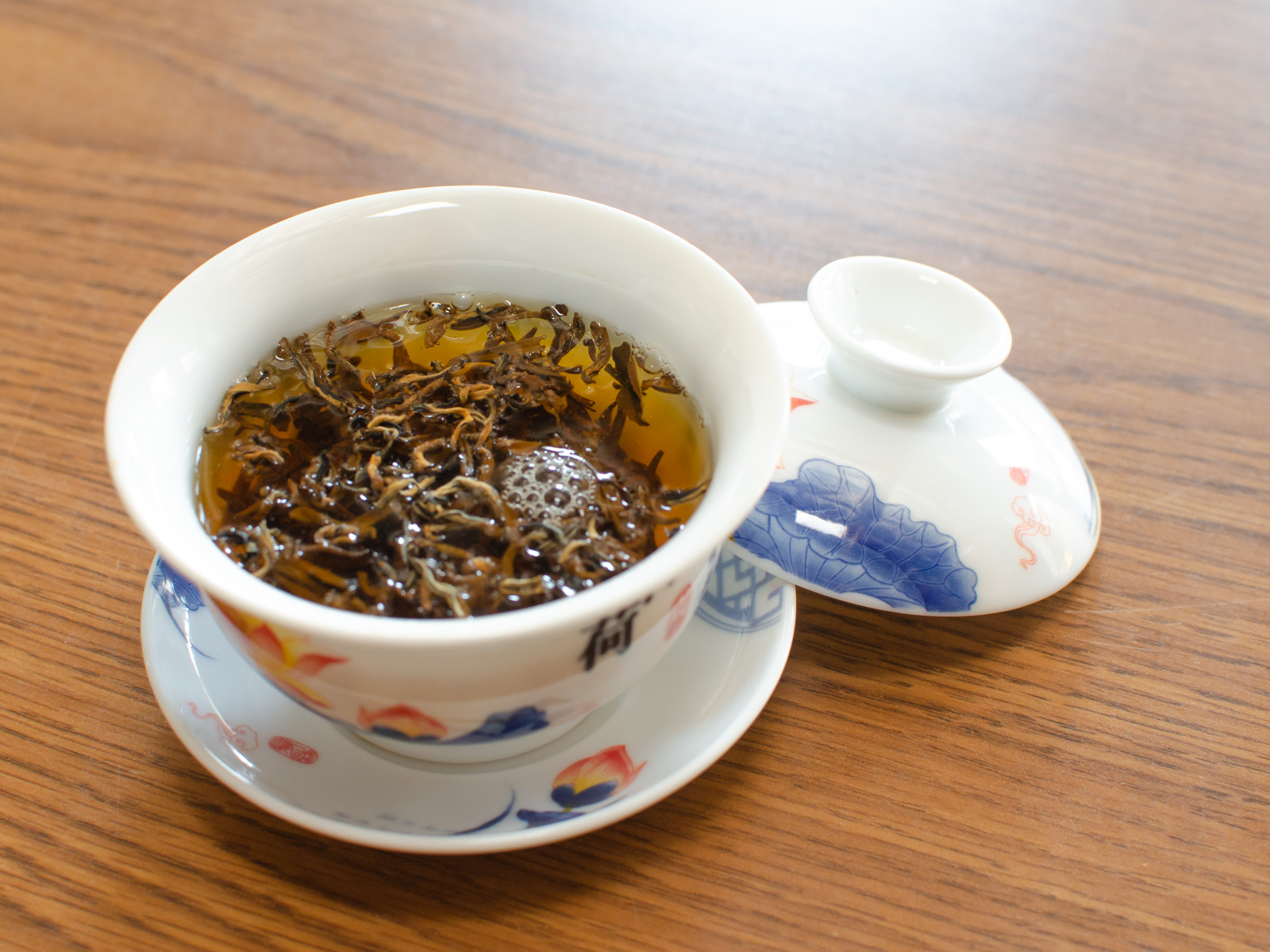 A delicate lotus-patterned gaiwan that’s just been filled with hot water - some of the Yunnan black tea leaves floating on top are still dry.