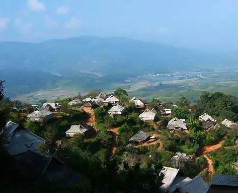 A cluster of low buildings among trees on a mountaintop, with a valley and another mountain beyond it in the distance.