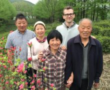 Five people stand together next to pinkish-red azalea bushes with green tea bushes and tall trees behind them.