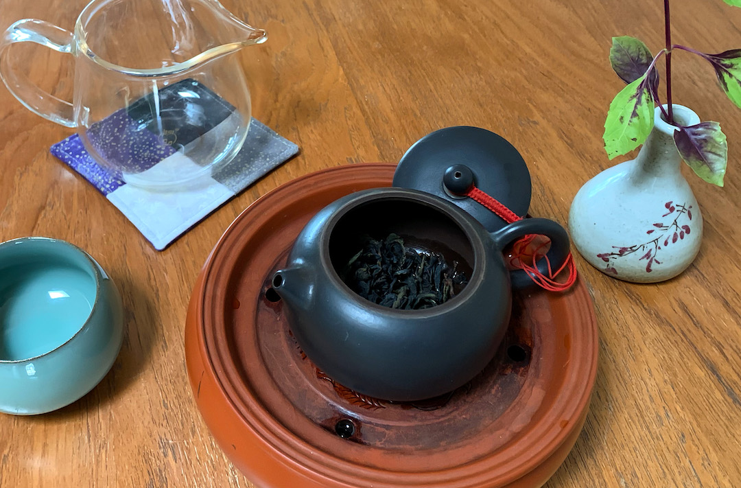 An open black yixing teapot filled with dry rock wulong tea leaves, set on a tray and surrounded by a pitcher, cup, and vase with a small plant.