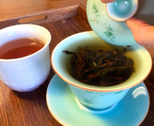 Looking closely at the black tea leaves of Zijuan Hong brewing in a pale blue-green gaiwan with its lid lifted ajar, accompanied by a full cup of tea on a wooden tray.