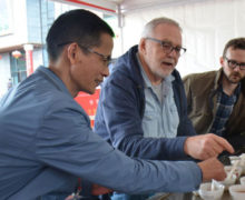 Three people smiling and talking while trying tea at a table in an outdoor tent.