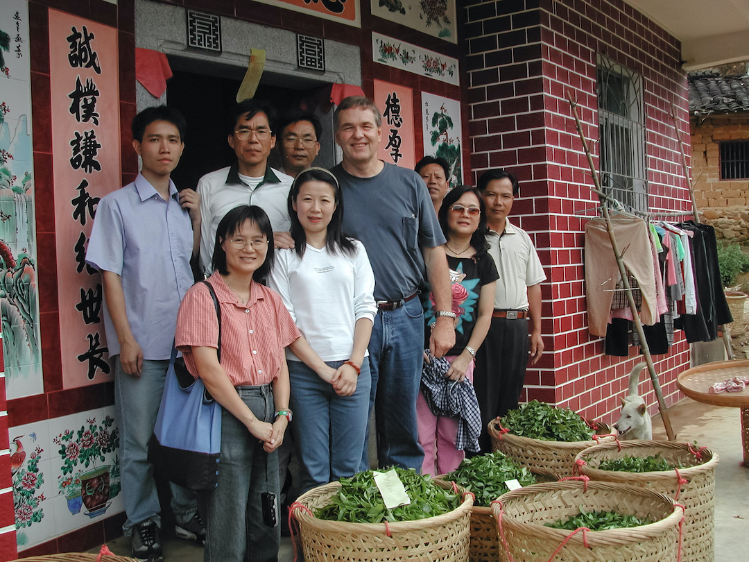 A group of people gathered in front of a building with large baskets of freshly harvested tea leaves in front of them.