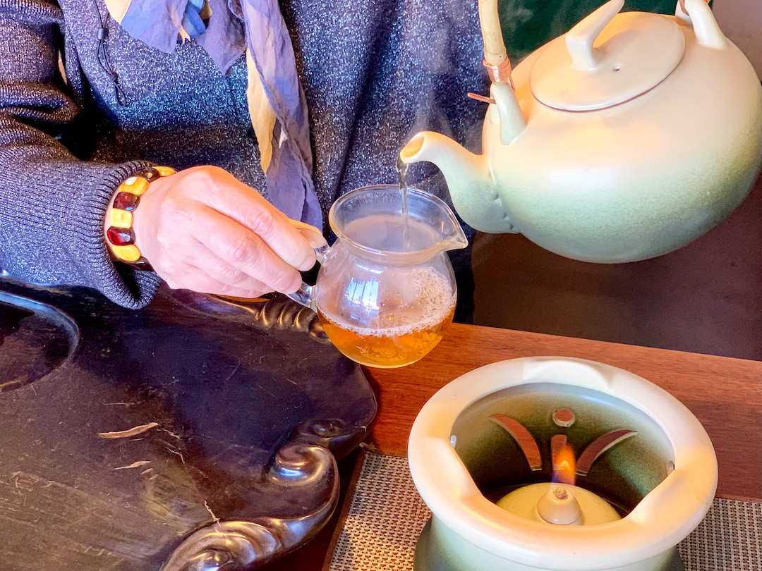 A hand holding a glass pitcher that dark golden puer tea is being poured into from a teapot, steaming hot.
